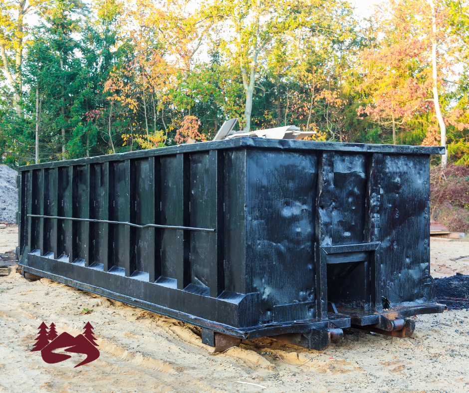 A black commercial roll-off dumpster. Fall trees are behind it.